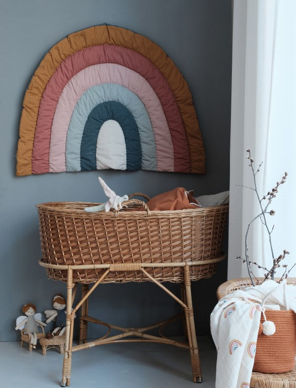 Rainbow play mat or wall hanging in muted colours displayed on wall above a Moses basket with dolls, curtain, plant and blanket on a chair.